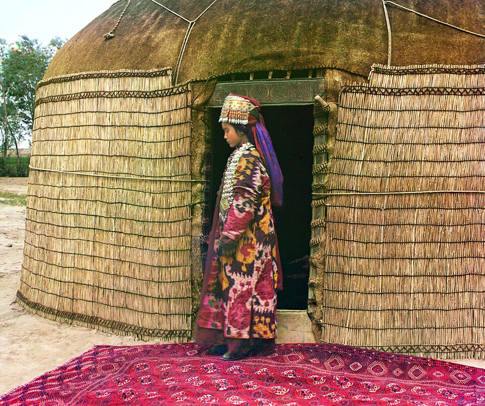Portrait en pied d'une femme de profil, debout sur un tapis à l'entrée d'une yourte, vêtue de vêtements et de bijoux traditionnels.