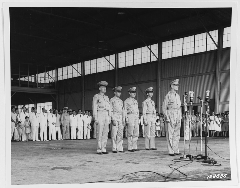 MacArthur, en uniforme, se tient devant quatre microphones sur pieds. Derrière lui, quatre hommes en uniforme militaire sont au garde-à-vous. Une foule nombreuse, composée d'hommes, de femmes et d'enfants élégamment vêtus de jupes, de tailleurs et d'uniformes, les observe.