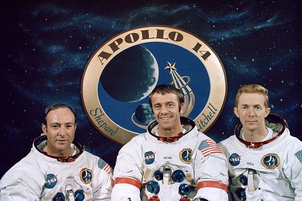 Mitchell, Shepard souriant et Roosa portent leurs combinaisons spatiales Apollo sans les casques. Leurs combinaisons portent leurs noms, l'écusson de la mission sur la poitrine gauche et l'écusson de la NASA sur la droite. Ils portent le drapeau américain sur leur manche gauche. Shepard a des anneaux rouges dans les bras. En arrière-plan se trouve un écusson de mission géant, entouré d'espace noir et d'étoiles.