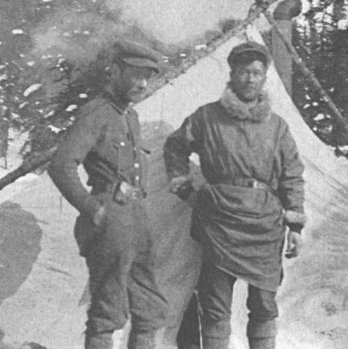 Sur une photographie en niveaux de gris, deux hommes se tiennent devant une tente et des conifères enneigés