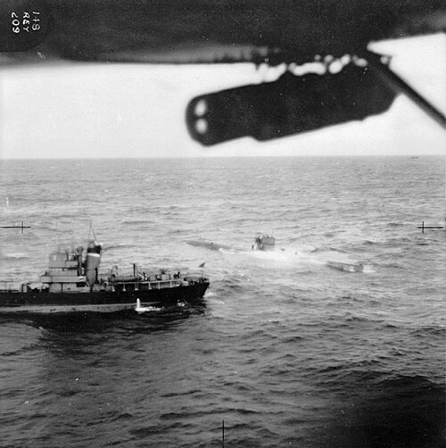 Photographie aérienne d'un sous-marin en surface dans une mer agitée ; un petit navire se trouve à proximité et les deux semblent immobiles. On peut voir une partie de l'aile gauche de l'avion, avec une bombe qui y est suspendue.