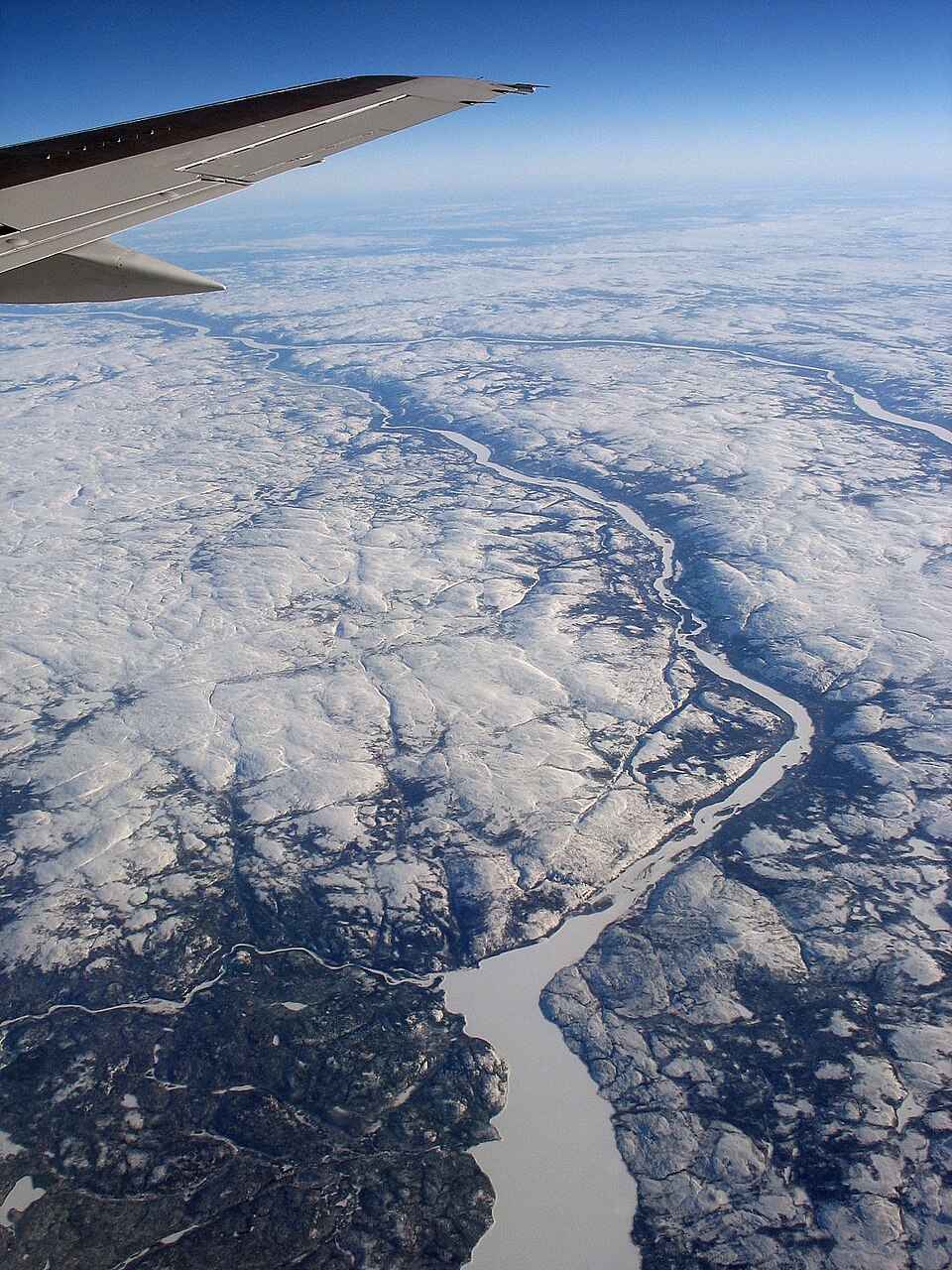 Photo aérienne prise en plongée, montrant des rivières. Le sol est enneigé, avec des arbres en bas à gauche et dans les vallées.