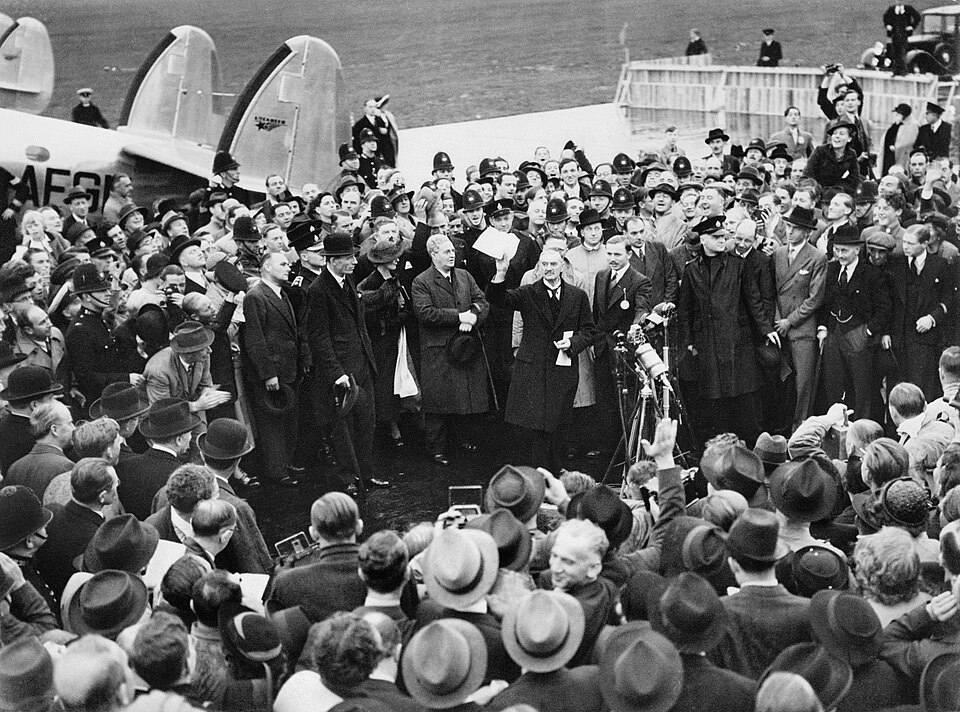 Une foule nombreuse sur un aérodrome ; le Premier ministre britannique Neville Chamberlain présente une lettre d'assurance du chancelier allemand Adolf Hitler.