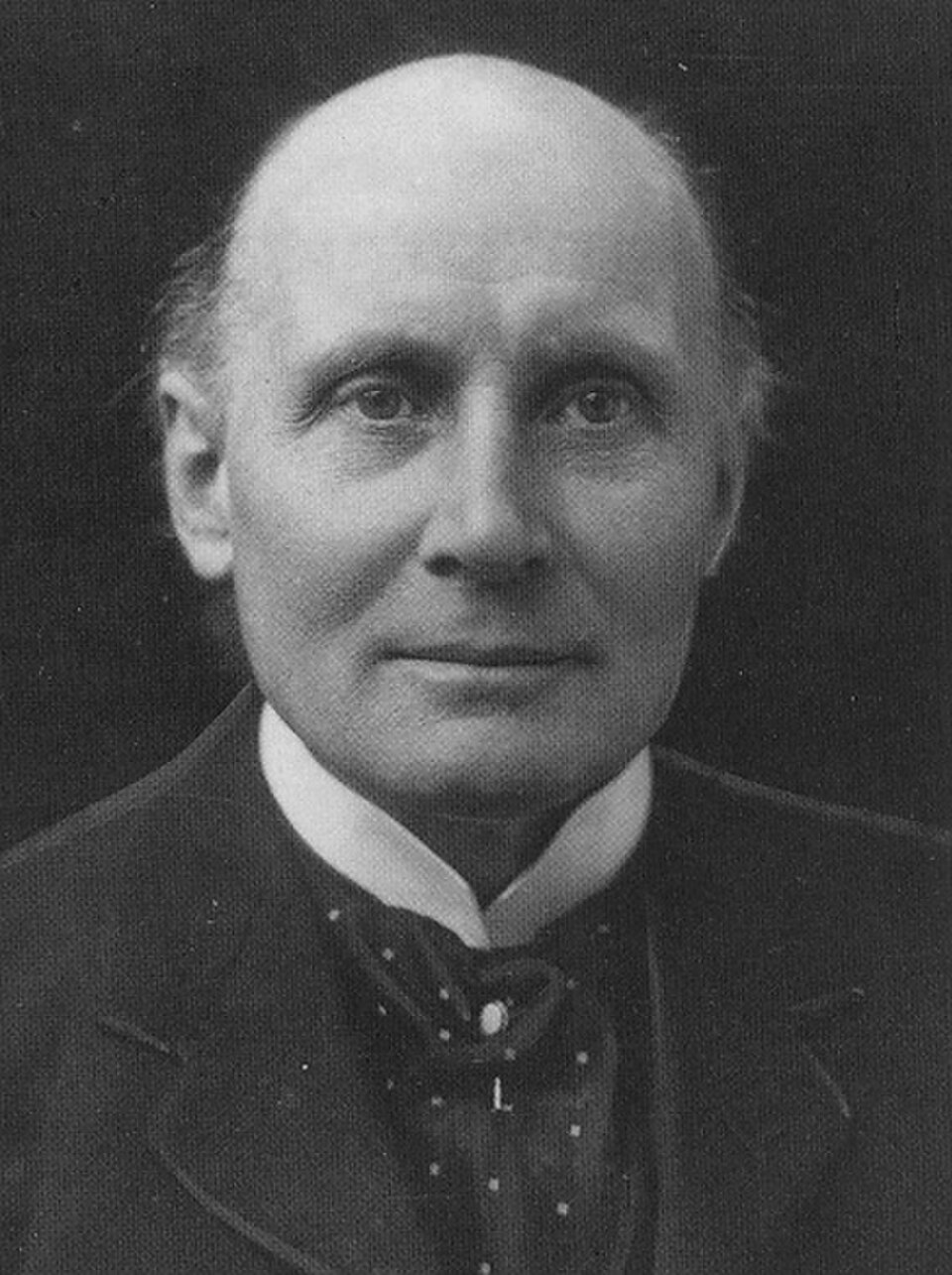 Photo montrant Alfred North Whitehead de face sur fond sombre, regardant l'objectif, vêtu d'un costume sombre et formel avec une chemise blanche à col montant en dessous