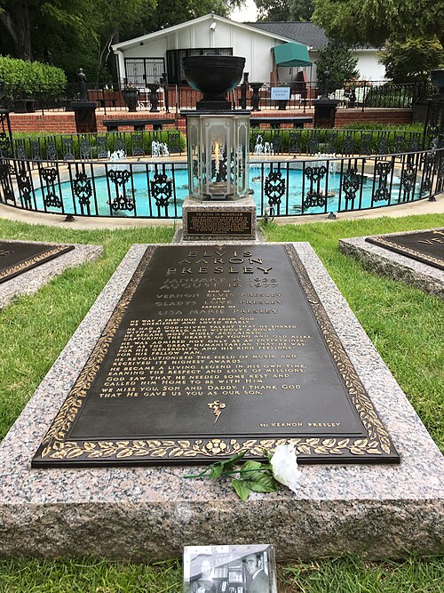 Une longue pierre tombale au niveau du sol porte l'inscription « Elvis Aaron Presley », suivie des dates de décès du chanteur, des noms de ses parents et de sa fille, ainsi que de plusieurs paragraphes de texte plus courts. À l'arrière-plan se trouve un petit bassin rond, avec une clôture métallique décorative basse et plusieurs fontaines.