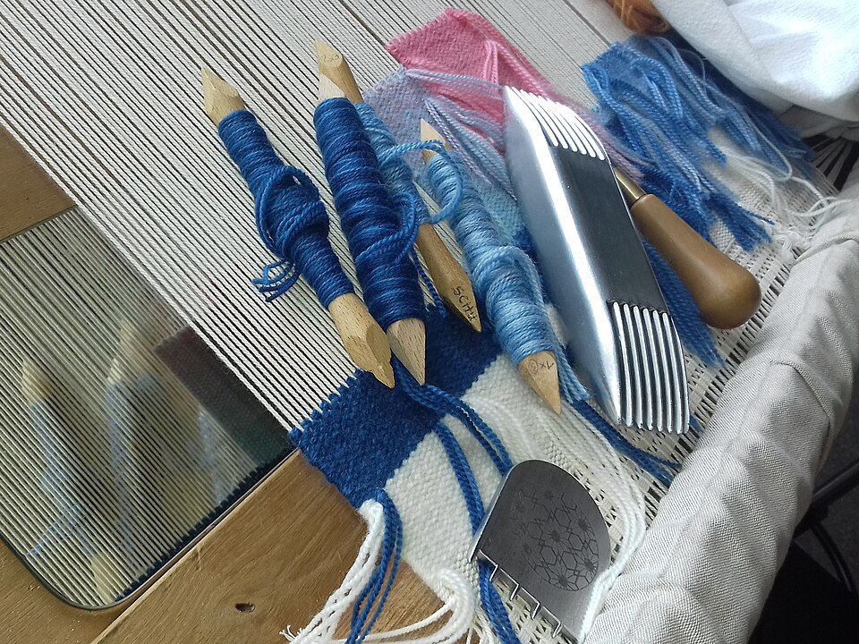 Outils de tapisserie sur un métier à tisser : miroir, os (enveloppés de fil), grattoir (à dents courtes), peigne lourd (à double extrémité, pour tendre la trame) et poinçon (pointe cachée).