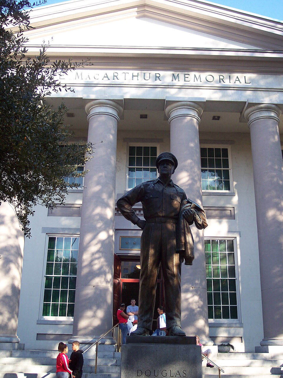 Une imposante statue en bronze de MacArthur se dresse sur un piédestal devant un grand bâtiment blanc à colonnes. Une inscription sur le bâtiment indique : « Mémorial Douglas MacArthur ».