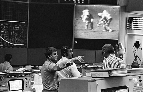 Une salle de contrôle ; visibles sur un grand écran, deux astronautes marchant sur la Lune