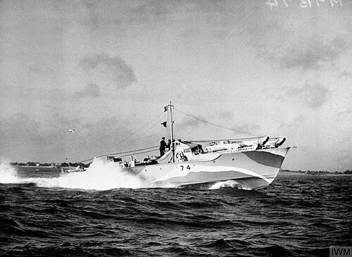 Navire filant à toute vitesse en mer avec une vague d'étrave blanche ; la terre est visible en arrière-plan