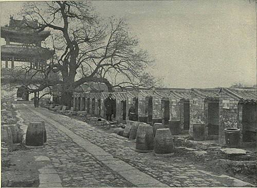 Une image en noir et blanc d'une allée pavée allant du bas à droite au haut à gauche menant à une porte à trois toits et bordée à droite par une rangée de petites cabines couvertes ouvertes sur un côté.