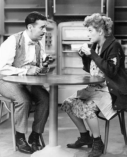 Deux personnages sont assis à table et discutent avec des expressions faciales expressives, une scène courante dans les sitcoms.