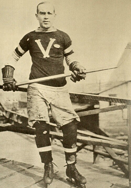 Un homme pose pour une photo portant des patins et un chandail de hockey sur glace, et tenant un bâton dans ses mains.