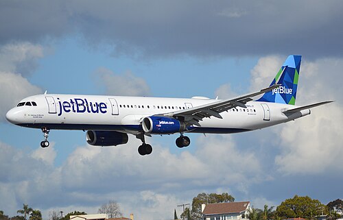 Un avion blanc avec les mots « jetBlue » peints à l'avant et une dérive bleu-vert s'approche de l'atterrissage avec son train d'atterrissage déployé alors qu'il plane au-dessus des maisons en contrebas