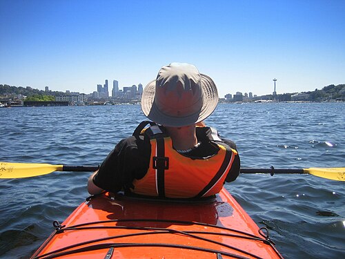 Photo de l'arrière d'une personne portant un gilet de sauvetage orange assis dans un kayak avec des bâtiments en arrière-plan