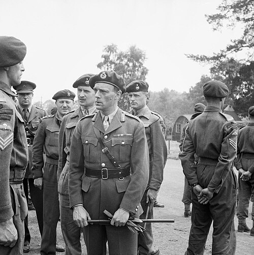 Officier général avec des gants et une canne à la main, parlant à un sergent, entouré d'autres officiers et hommes