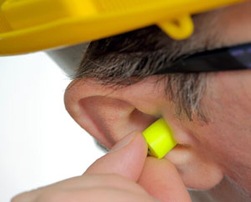 Une image de la tête d'un homme, focalisée sur l'oreille, avec des bouchons d'oreille jaunes insérés dans cette oreille.