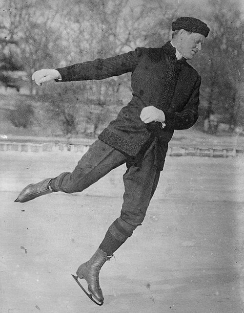 Homme d'une trentaine d'années, portant un pantalon et un manteau foncés, patinant sur une patinoire extérieure, tourné vers la droite et regardant vers le bas en direction de la glace